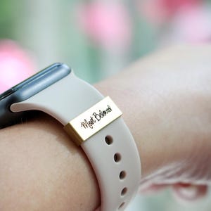 Może przedstawiać: Zbliżenie smartwatcha z jasnobeżowym paskiem i złotym metalowym akcentem. Akcent zawiera odręczny tekst "Most Beloved". Pasek zegarka ma kilka otwor&oacute;w do regulacji.