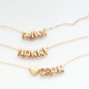 May include: Three gold-colored necklaces with pendant names: "MIMI", "NANNY", and "GRAN". The "GRAN" necklace features a heart-shaped pendant. Each necklace has a delicate chain and is displayed against a white background.