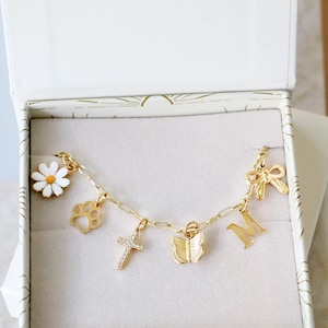 Op de afbeelding: Een goudkleurige bedelarmband met een witte margriet, een pootafdruk, een met kristallen ingelegd kruis, een vlinder, de letter M en een strik, gepresenteerd in een witte doos met de tekst Hello Beautiful Be yourself with some extra shine. You're a gem xx.