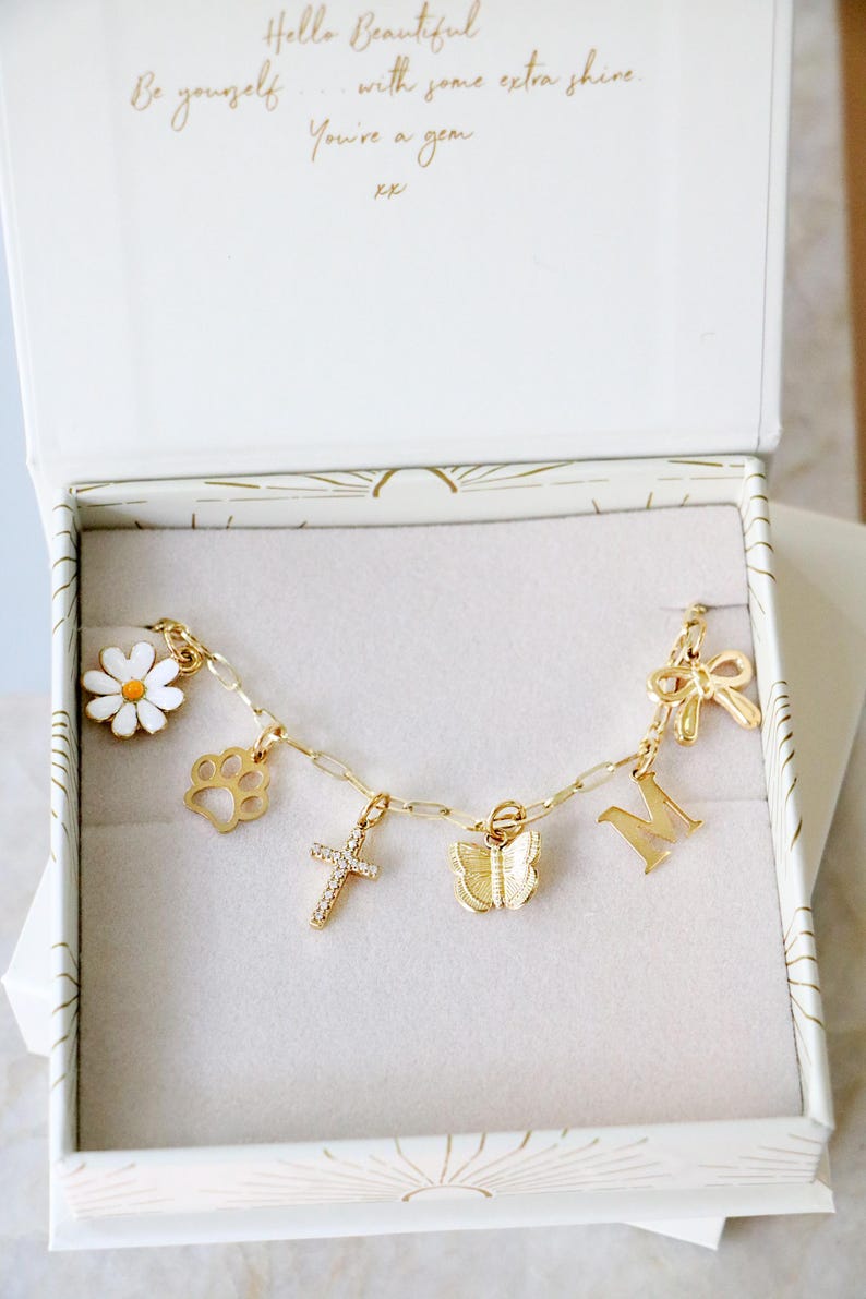 Op de afbeelding: Een goudkleurige bedelarmband met een witte margriet, een pootafdruk, een met kristallen ingelegd kruis, een vlinder, de letter M en een strik, gepresenteerd in een witte doos met de tekst Hello Beautiful Be yourself with some extra shine. You're a gem xx.