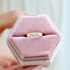 May include: A gold band ring featuring the engraved cursive text "miller" on its outer surface, securely positioned within a hexagonal, dusty pink velvet ring box with a central slot for holding the jewelry piece in place.