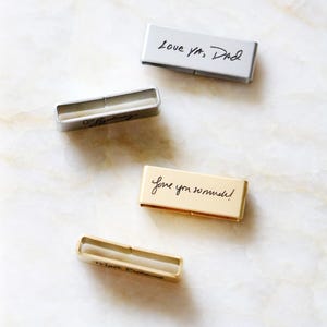 Może przedstawiać: Cztery prostokątne metalowe pendrive'y z odręcznymi wiadomościami. Pendrive'y są w odcieniach srebra i złota. Wiadomości obejmują "Love ya, Dad" i "Love you so much!"