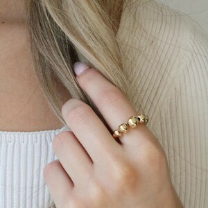 May include: A gold ring featuring a series of small, evenly spaced spheres. The ring is worn on a finger, with the hand and arm visible. The ring's design is simple and elegant, suitable for everyday wear.