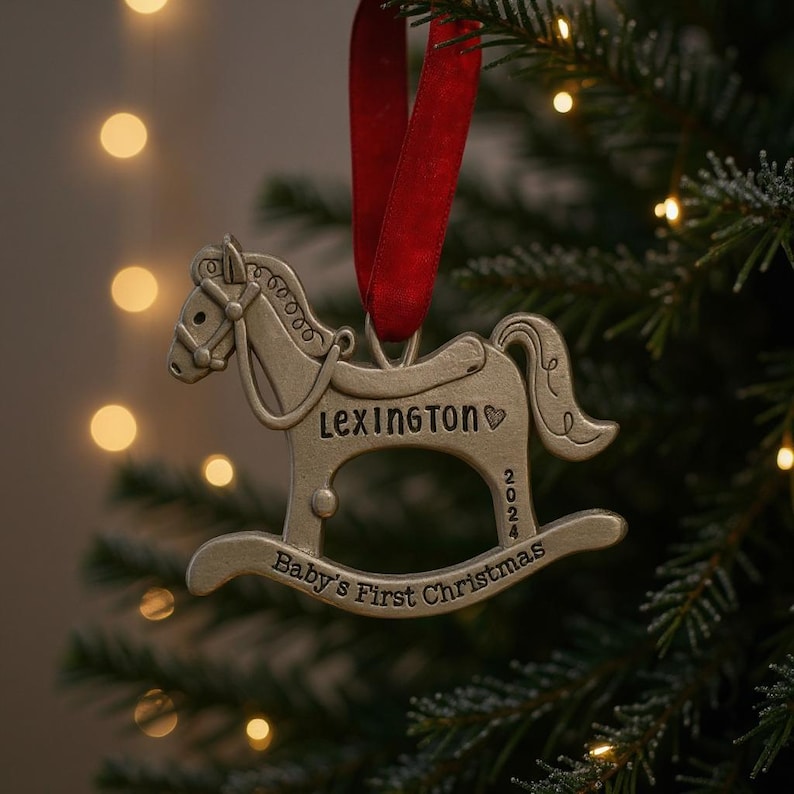 以下が含まれることがあります： 「Lexington」と「Baby's First Christmas 2024」の文字が入った、銀色のロッキングホースのクリスマスオーナメント。赤いリボンで吊るされ、クリスマスツリーを背景にしています。