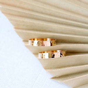 May include: Two gold hammered wedding bands on a beige textured background.