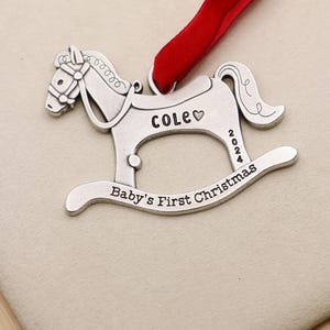 以下が含まれることがあります： 赤いリボンが付いた、銀色のロッキングホースのクリスマスオーナメント。「COLE」という名前とハート、「2024」の年号、「Baby's First Christmas」という文字が刻まれています。