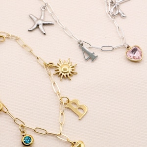 Op de afbeelding: Twee bedelarmbanden met ovale schakels. De zilverkleurige armband heeft een blauw boze oog, een zeester, een strik, het cijfer 4 en een roze hartje. De goudkleurige armband heeft een zon, de letter B, een blauwe steen en een hartje.