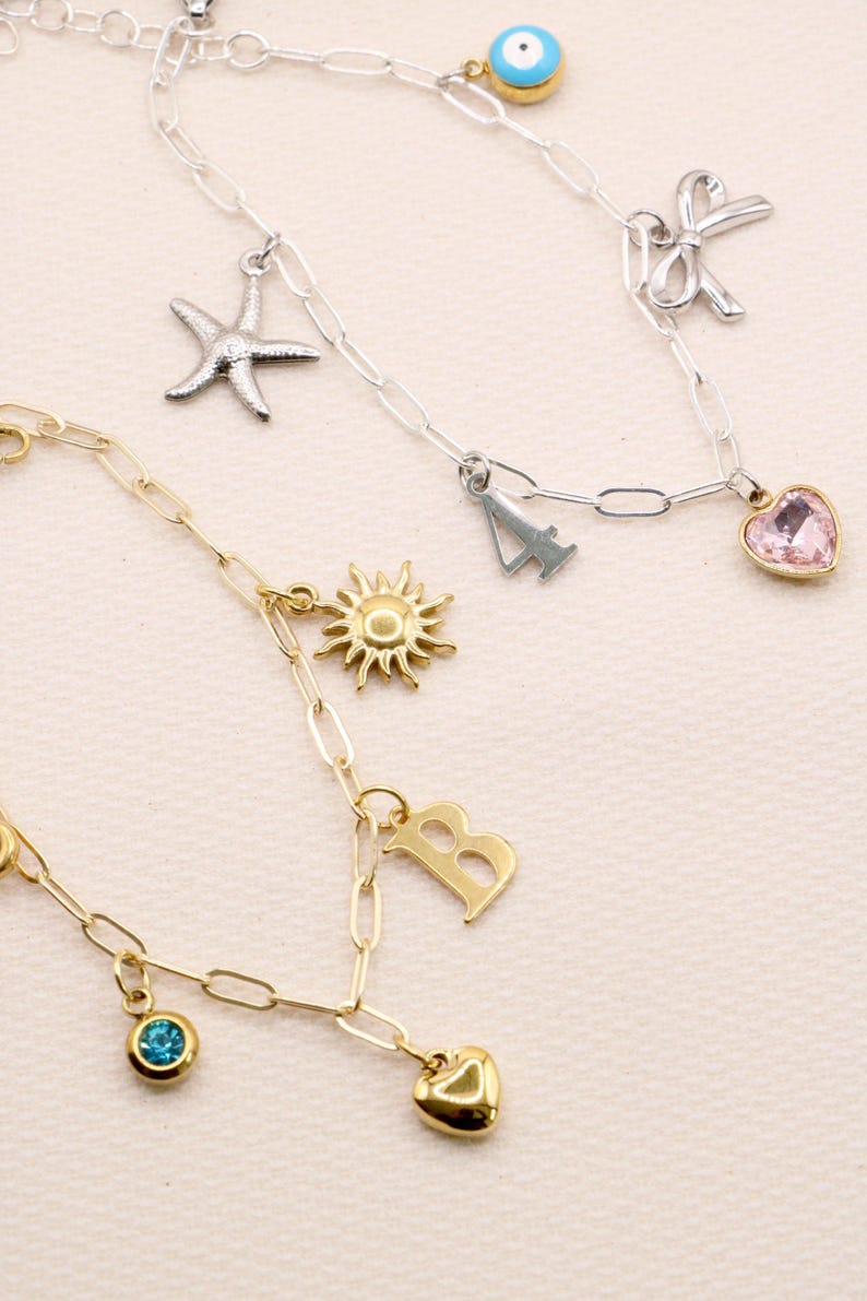 Op de afbeelding: Twee bedelarmbanden met ovale schakels. De zilverkleurige armband heeft een blauw boze oog, een zeester, een strik, het cijfer 4 en een roze hartje. De goudkleurige armband heeft een zon, de letter B, een blauwe steen en een hartje.