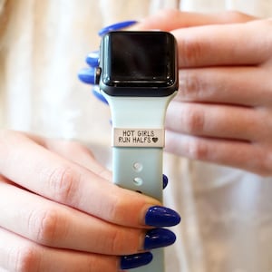 Może przedstawiać: Czarny smartwatch z jasnoniebieskim paskiem. Pasek ma wygrawerowany napis "HOT GIRLS RUN HALFS" na srebrnej metalowej płytce. Zegarek trzyma osoba z niebieskim lakierem do paznokci.