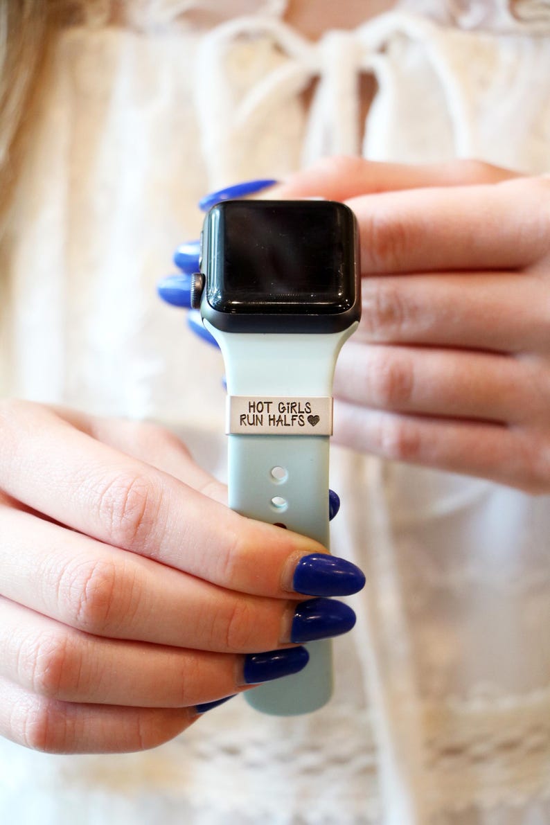 Może przedstawiać: Czarny smartwatch z jasnoniebieskim paskiem. Pasek ma wygrawerowany napis "HOT GIRLS RUN HALFS" na srebrnej metalowej płytce. Zegarek trzyma osoba z niebieskim lakierem do paznokci.