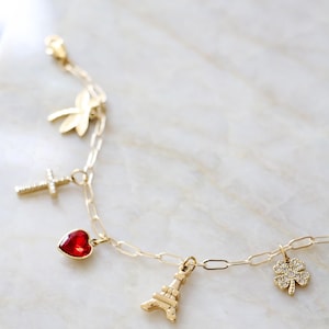 Op de afbeelding: Een goudkleurige kettingarmband met vijf verschillende bedels: een gouden libel, een kruis met heldere steentjes, een rode hartvormige steen, een gouden Eiffeltoren en een klavertjevier met heldere steentjes.