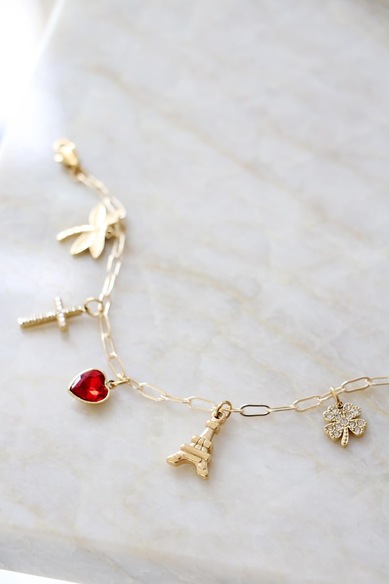 Op de afbeelding: Een goudkleurige kettingarmband met vijf verschillende bedels: een gouden libel, een kruis met heldere steentjes, een rode hartvormige steen, een gouden Eiffeltoren en een klavertjevier met heldere steentjes.