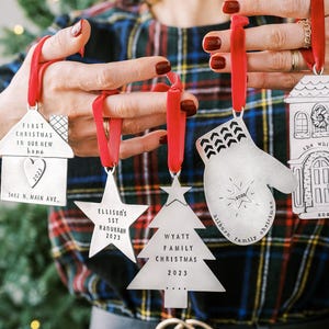 以下が含まれることがあります： 赤いリボンが付いた銀色のクリスマスオーナメント。オーナメントには、家、星、ミトン、木、家のデザインが含まれています。各オーナメントには、「最初のクリスマス」や「家族のクリスマス」などの名前、日付、フレーズなどのテキストが記載されています。