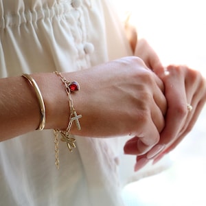 Op de afbeelding: Een goudkleurige schakelarmband met drie bedels: een rode hartvormige steen, een klein kruisje bezet met doorzichtige steentjes en een goudkleurige libel. De armband wordt gedragen naast een eenvoudige, gladde goudkleurige armband.