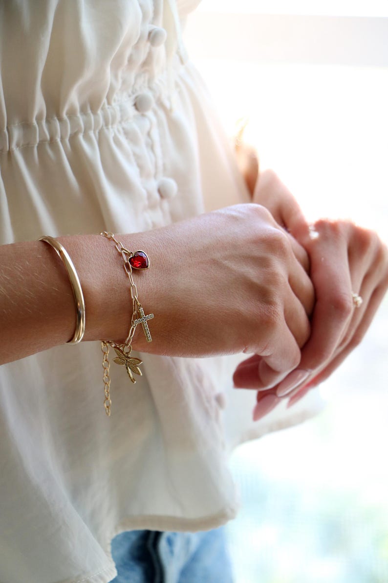 Op de afbeelding: Een goudkleurige schakelarmband met drie bedels: een rode hartvormige steen, een klein kruisje bezet met doorzichtige steentjes en een goudkleurige libel. De armband wordt gedragen naast een eenvoudige, gladde goudkleurige armband.