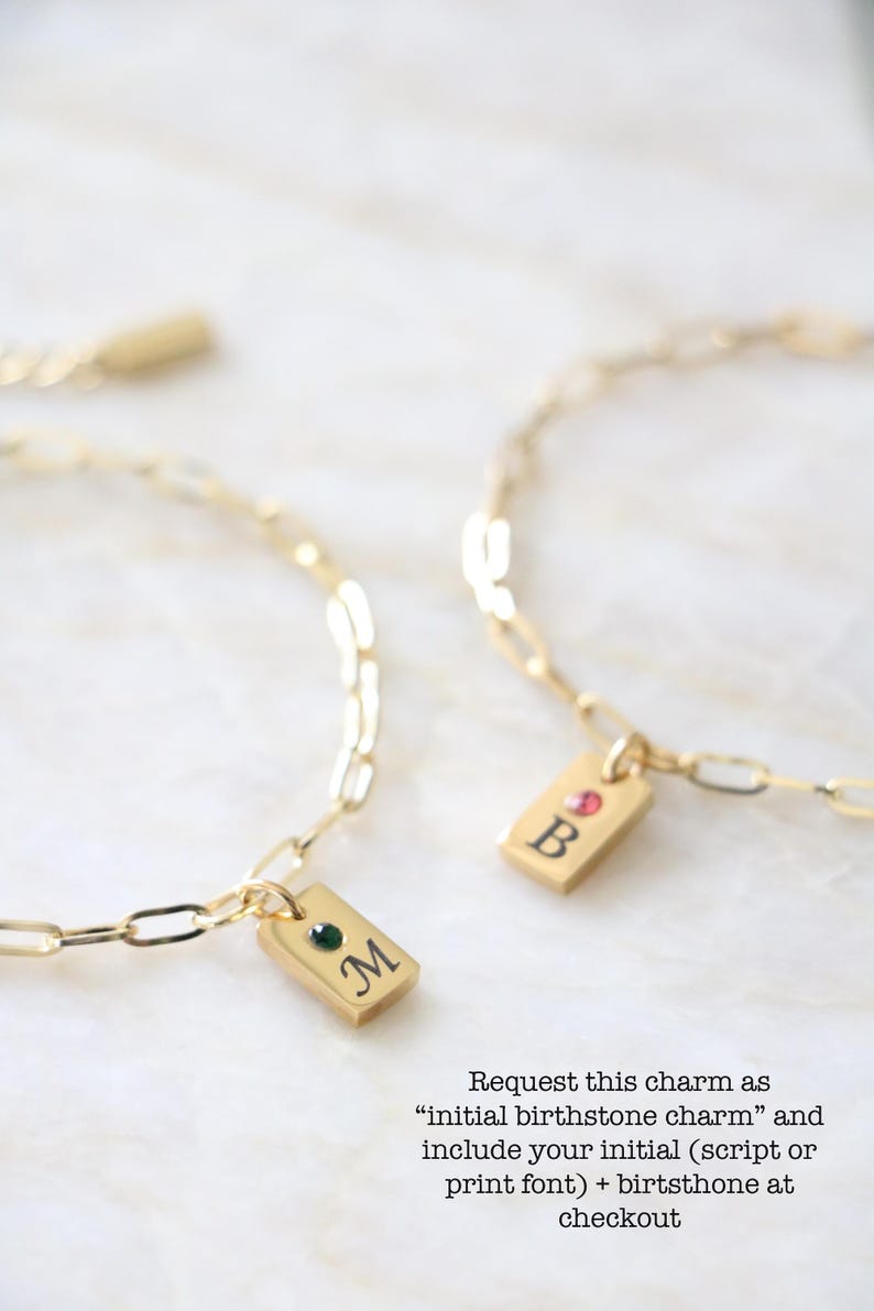 Op de afbeelding: Twee goudkleurige kettingarmbanden met rechthoekige bedels. E&eacute;n bedel toont de letter M met een groene steen, de andere de letter B met een roze steen. De tekst luidt: Request this charm as initial birthstone charm and include your initial (script or print font) + birthstone at checkout.