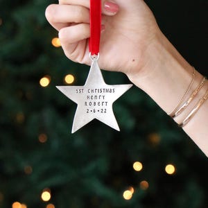以下が含まれることがあります： 「1st Christmas Henry Robert 2.6.22」の文字が刻まれた、銀色の星型のクリスマスオーナメント。赤いベルベットのリボンから吊り下げられています。背景は、ぼやけたクリスマスツリーとボケライトです。