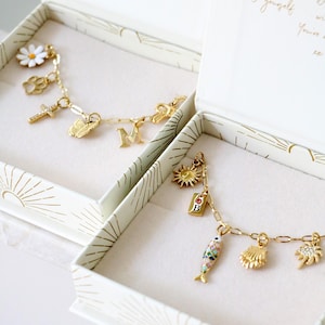Op de afbeelding: Twee goudkleurige bedelarmbanden in open witte doosjes met zonnestraalpatronen. De ene armband heeft een madeliefje, pootafdruk, kruis, vlinder en letter M. De andere bevat een zon, een B-label, een vis, een schelp en een palmboombedel.