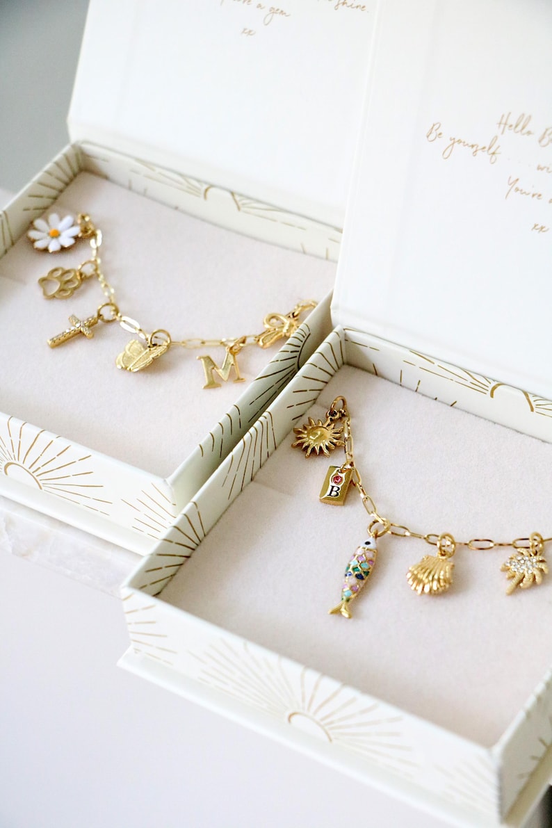 Op de afbeelding: Twee goudkleurige bedelarmbanden in open witte doosjes met zonnestraalpatronen. De ene armband heeft een madeliefje, pootafdruk, kruis, vlinder en letter M. De andere bevat een zon, een B-label, een vis, een schelp en een palmboombedel.