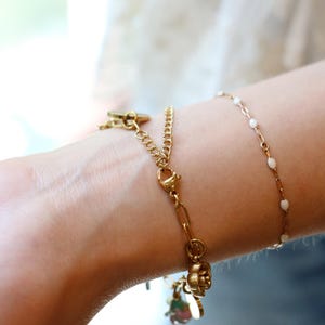 Op de afbeelding: Twee goudkleurige armbanden die samen worden gedragen. De ene is een dikkere schakelarmband met een karabijnsluiting en een kleine bloembedel. De andere is een fijne, dunne ketting met kleine, gelijkmatig verdeelde witte emaille kraaltjes.
