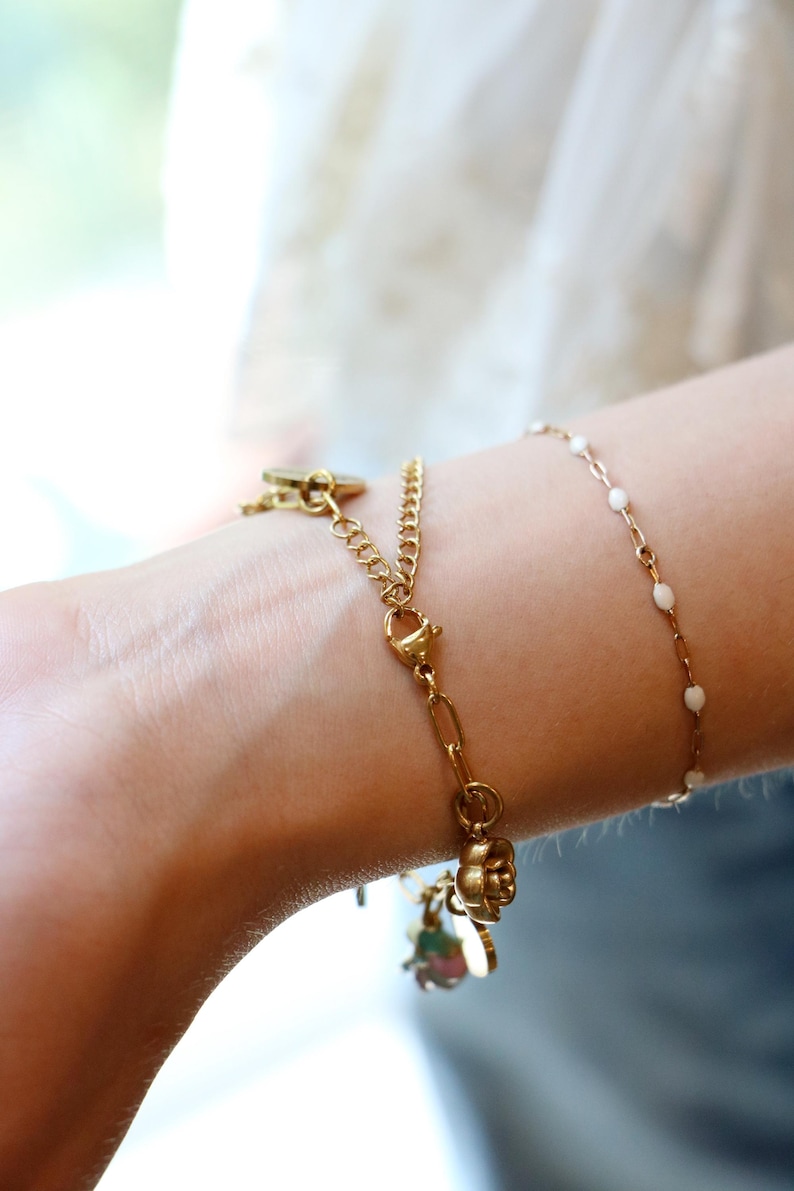 Op de afbeelding: Twee goudkleurige armbanden die samen worden gedragen. De ene is een dikkere schakelarmband met een karabijnsluiting en een kleine bloembedel. De andere is een fijne, dunne ketting met kleine, gelijkmatig verdeelde witte emaille kraaltjes.
