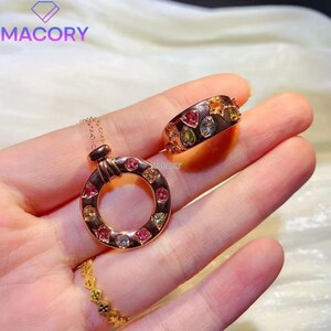 May include: A rose gold pendant necklace featuring a circular charm set with multi-colored gemstones, a matching wide band ring with inlaid colorful stones, and a thin gold ring with a textured, repeating geometric pattern.