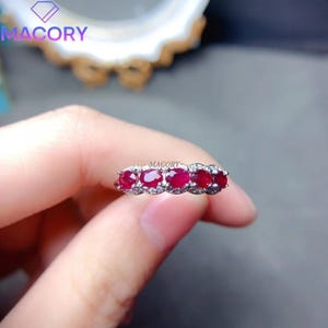 May include: A silver-toned ring featuring five oval-cut deep red rubies arranged in a horizontal row, accented by small clear stones set along the band. The word MA CORY is printed in small text above the center of the ruby gemstone setting.
