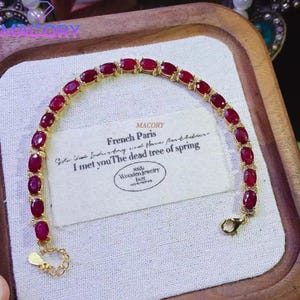 May include: A gold-toned bracelet featuring a series of oval-shaped, deep red gemstones linked together. The bracelet includes a lobster claw clasp and an adjustable chain extension. The piece is displayed on a white fabric card with printed text.