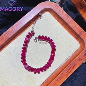 May include: A silver-toned bracelet featuring a continuous row of oval-shaped, deep red gemstones set in metal prongs. The piece includes a delicate chain extension with a lobster claw clasp and the text MACORY printed on the display surface.