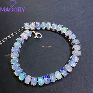 May include: A silver-toned bracelet featuring a continuous row of oval-shaped, iridescent opals with blue and green flashes. The jewellery piece includes a metal chain extension and a lobster claw clasp. The text MACORY is visible in the image.