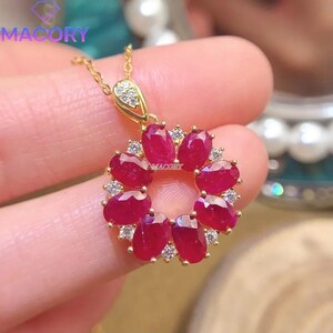 May include: A gold pendant necklace featuring a circular arrangement of eight oval-cut deep red rubies interspersed with small clear white stones. The pendant includes a triangular bail set with tiny white stones and a delicate gold chain.