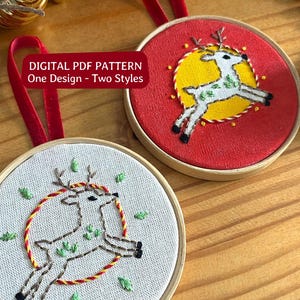 Pode incluir: Dois bastidores de bordado circulares de madeira com um design de rena. Um tem um fundo de tecido vermelho com um círculo amarelo, o outro um fundo cinza claro. Ambos incluem fitas de veludo vermelho e o texto: DIGITAL PDF PATTERN One Design - Two Styles.