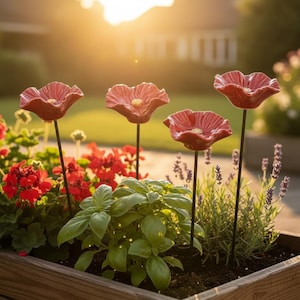 May include: Four red ceramic poppy flower garden stakes with thin black metal stems, positioned upright in a wooden planter box filled with green basil, lavender, and red geraniums, illuminated by warm golden hour sunlight.