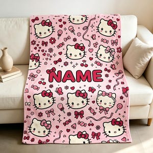 May include: A light pink fleece blanket featuring a repeating pattern of Hello Kitty faces, red bows, cherries, lollipops, and hearts. The word NAME is printed in large, bold, dark pink capital letters across the centre of the blanket.