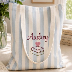 Op de afbeelding: Een lichtblauw en wit gestreepte canvas tas met de geborduurde naam Audrey in donkerrood schrift, een klein roze hartje en een stapel van twee boeken met groene en rode kaften in het midden, voorzien van twee lange witte schouderbanden.