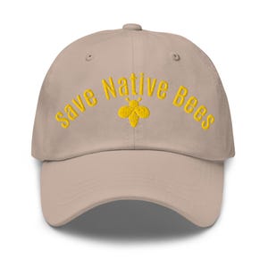 May include: A beige baseball cap featuring the embroidered text "Save Native Bees" in yellow thread, arched over a small, stylized yellow bee icon centered on the front panel. The cap has a structured crown and a curved brim.