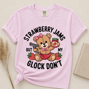 May include: A light pink t-shirt featuring a graphic of a brown teddy bear wearing a pink bow and skirt. The bear holds a handgun and a jar of strawberry jam. The text reads: STRAWBERRY JAMS BUT MY GLOCK DON'T. The shirt has a crew neckline.