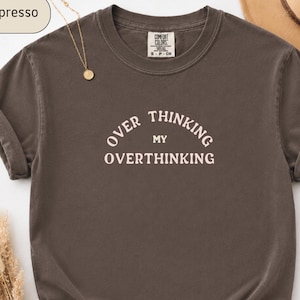 May include: A dark brown short-sleeve t-shirt with a crew neckline and rolled sleeves. The centre of the chest features the cream-coloured text "OVER THINKING MY OVERTHINKING" arranged in an arched layout. The shirt has a Comfort Colors brand label.