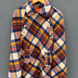 May include: A double-breasted cape coat featuring a plaid pattern in shades of orange, navy blue, red, and cream. The garment has a wide collar, six round buttons arranged in two vertical rows, and two rectangular front pockets.