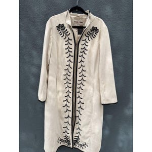 May include: A long, cream-colored coat featuring a full-length front zipper and black embroidered leaf-like patterns running vertically along the opening. The coat has long sleeves with black trim at the cuffs and a high collar, with the label Powder River.