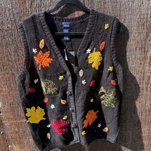 May include: A black knit button-down vest featuring embroidered autumn leaf motifs in shades of orange, yellow, red, and green, along with small white flower accents. The garment has a V-neckline, ribbed trim, and a brand label at the inner collar.