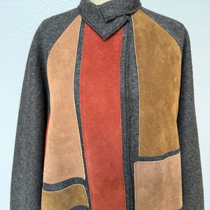 May include: A charcoal gray wool jacket featuring a high collar and asymmetrical panels of suede in shades of rust, tan, and brown. The garment has long sleeves and a structured front closure, showcasing a mix of textures and earth-toned colors.