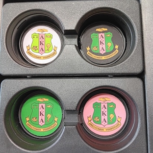 May include: A set of four circular car cup holder coasters featuring the Alpha Kappa Alpha sorority crest. The coasters include one white, one black, one green, and one pink background, each displaying the Greek letters A.K.A. and the motto.