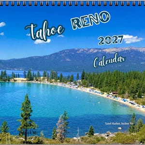 May include: A 2027 wall calendar featuring a scenic photograph of Sand Harbor in Incline Village, Nevada. The image displays a bright blue lake, a sandy beach with people, and dense pine forests under a clear sky. Text reads Tahoe RENO 2027 Calendar.