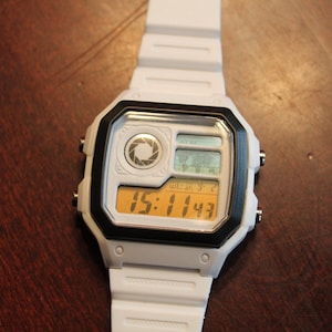 Könnte beinhalten: Eine weiße Digitalarmbanduhr mit schwarzer rechteckiger Lünette. Das Zifferblatt zeigt ein kreisförmiges Blendenlogo, eine kleine Weltkartengrafik und eine Digitalanzeige, die die Uhrzeit 15:11:43 und das Datum Mittwoch 9-2 in Gelb anzeigt.