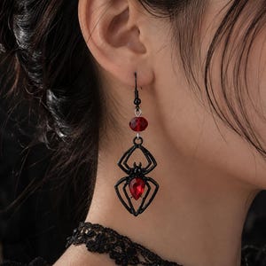 May include: A dangling earring featuring a black metal spider charm with a faceted red teardrop gemstone set in its abdomen. Above the spider, a small red bead is attached to a black metal hook, creating a cohesive dark and red color scheme.