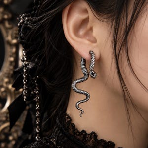 Puede incluir: Un pendiente de serpiente en tono plateado con un patrón texturizado de escamas en el cuerpo y una piedra negra incrustada en la cabeza. El diseño de la serpiente se curva hacia abajo desde el lóbulo, creando una silueta larga y colgante.