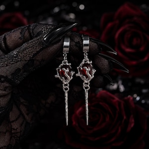 May include: A pair of silver-toned drop earrings featuring a deep red, teardrop-shaped stone encased in a textured, organic metal frame. Each earring hangs from a simple silver hoop and terminates in a long, slender, pointed metal spike.