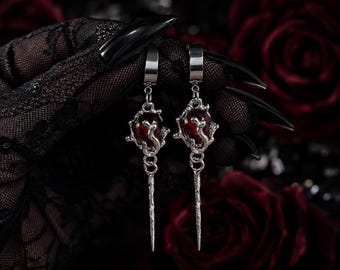 May include: A pair of silver-toned drop earrings featuring a deep red, teardrop-shaped stone encased in a textured, organic metal frame. Each earring hangs from a simple silver hoop and terminates in a long, slender, pointed metal spike.