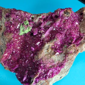 May include: A raw mineral specimen featuring a cluster of vibrant magenta-colored crystals embedded within a rough, tan-colored rock matrix. A small patch of bright lime green mineral deposits is visible on the upper left side of the crystal formation.